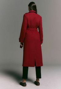 Red belted wool coat with a notched collar, two buttons on sleeves, and a vent at the back; paired with black pants and animal print flats.