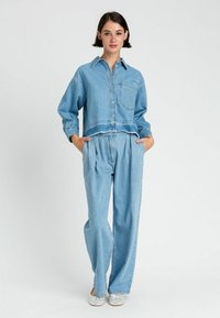 Completo in denim blu chiaro composto da una camicia corta con bottoni e tasca sul petto, abbinata a pantaloni larghi con pieghe e scarpe slip-on.