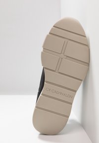 Basket noire avec un dessus en cuir texturé et une semelle en caoutchouc beige, présentant des rainures horizontales et "CK CALVIN KLEIN" embossé sur le côté.