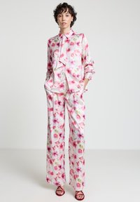 Seidenbluse mit Blumenmuster und Halsband, kombiniert mit weit geschnittenen Hosen in Rosa- und Grüntönen. Das Modell steht mit den Händen in den Taschen.