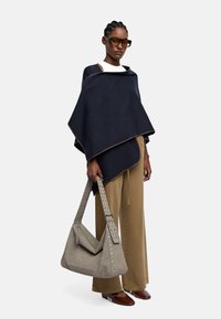 Granatowy wełniany poncho z kontrastowym wykończeniem, zestawione z jasnobrązowymi spodniami o szerokich nogawkach oraz szarym skórzanym torebką z ćwiekowymi akcentami.