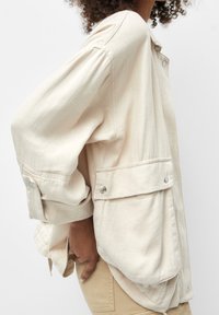 Chhemise beige à manches longues avec une coupe décontractée, dotée d'une poche avant, d'accents de boutons et d'une texture de tissu douce.