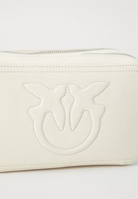 Borsa in pelle bianca con doppia cerniera e logo inciso di due uccelli centrato sul pannello anteriore su uno sfondo semplice.
