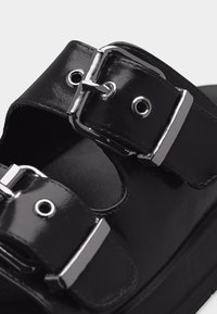 Μαύρο δερμάτινο σανδάλι με δύο ρυθμιζόμενες ασημένιες μεταλλικές αγκράφες και οπές, κοντινή λήψη που δείχνει τους υφασμένους ιμάντες και τη σόλα.