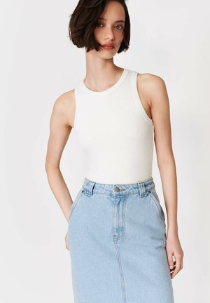 Lühikeste tumedate juustega naine, kes kannab varrukateta valget topi ja kõrge vöökohaga helesinist teksaseelikut, seisab valge tausta ees.