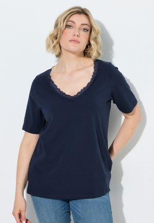 Vrouw met kort blond haar die een marineblauw T-shirt met V-hals, kanten randje en korte mouwen draagt, en een blauwe spijkerbroek, staand tegen een witte achtergrond.