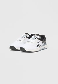 Pari di scarpe da atletica Reebok Nano XV bianche e nere con lacci, suole testurizzate e linguetta con marchio su uno sfondo grigio chiaro e semplice.