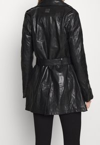 Czarna skórzana kurtka z paskiem w talii, długimi rękawami i kołnierzem. Ma na plecach metkę z logo oraz błyszczące, teksturowane wykończenie.