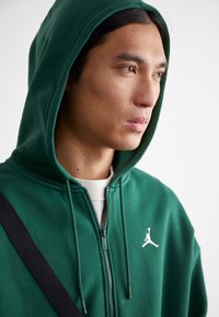 Grøn zip-hoodie lavet af blødt materiale, med hætte med snøre og et hvidt logo på brystet, parret med en lyse skjorte under.