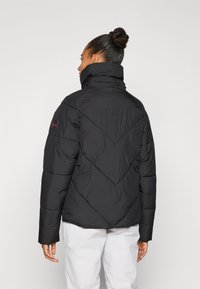 Doudoune noire avec un col haut, des motifs de quilting verticaux et diagonaux, et un logo rond sur l'épaule gauche.