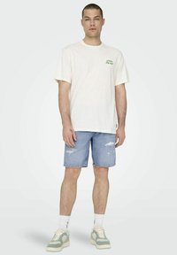 Camiseta de algodón blanca con estampado "Persigue el sol" en verde, combinada con pantalones cortos de mezclilla azul claro con detalles desgastados. Calcetines blancos y zapatillas.
