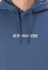 Námornícky modrá mikina s kapucňou vyrobená z jemného materiálu, s prednou klokankou a šnúrkami. Zobrazí logo "Hollister" v bielej farbe.