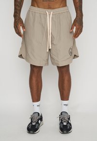Shorts beige con cintura elástica y cordón, con aberturas laterales y un logo en el dobladillo. Llevados con zapatillas negras y grises.