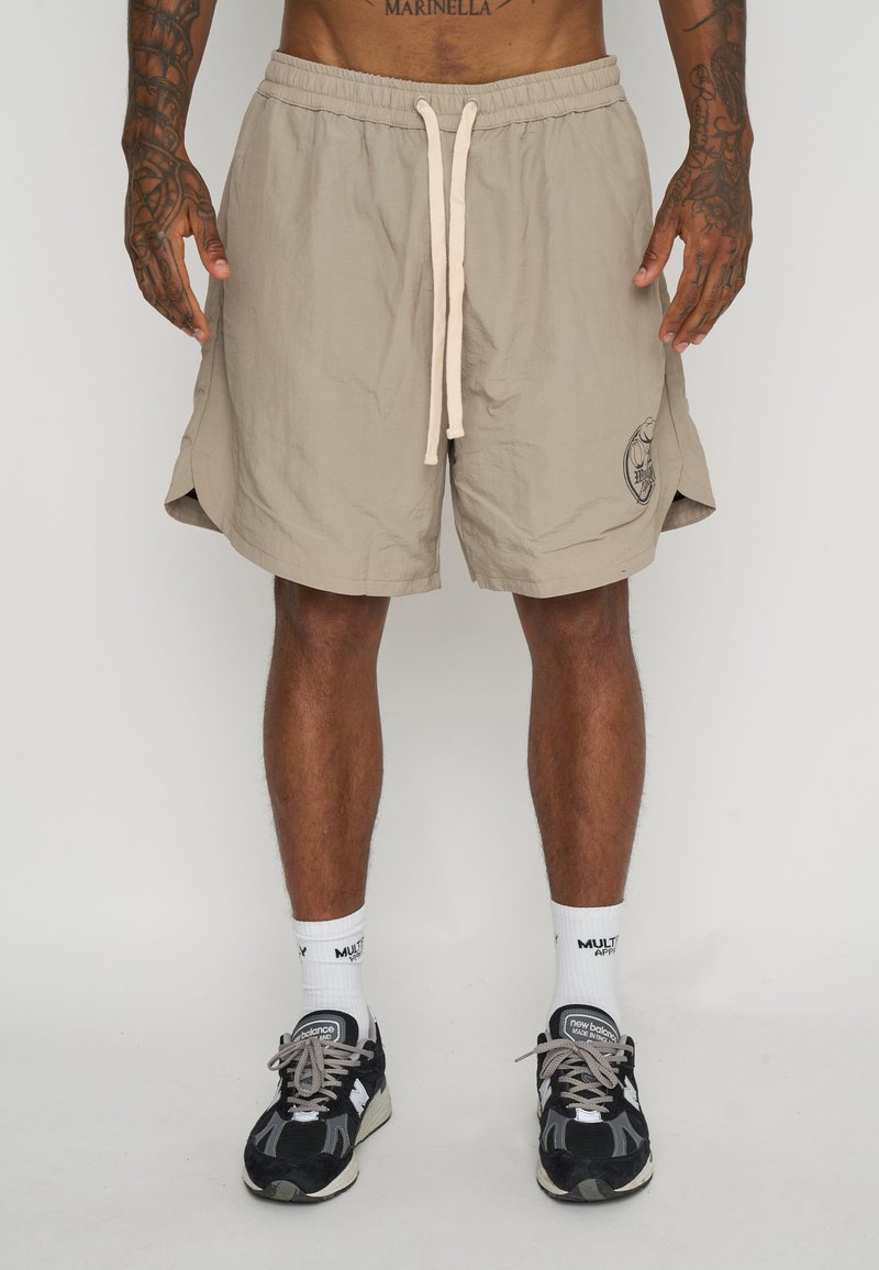 Shorts beige con cintura elástica y cordón, con aberturas laterales y un logo en el dobladillo. Llevados con zapatillas negras y grises.