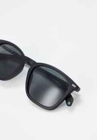 Ochelari de soare negri cu rame groase, lentile polarizate și un finisaj mat. Prezintă accente argintii pe brațe și un model geometric pe lateral.