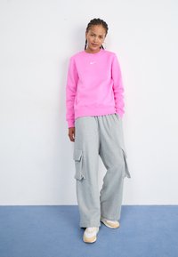 Sweatshirt rosa intenso com decote redondo e logotipo branco da Nike, combinado com calças cargo cinzas soltas com bolsos laterais e sapatilhas bege.