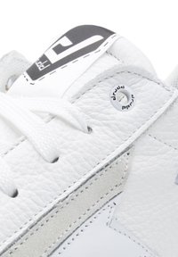 Białe skórzane trampki z teksturowanym wierzchem, płaskimi białymi sznurówkami, szarymi zamszowymi akcentami i czarnym logo na języku. Posiadają wzmocnioną oczko.