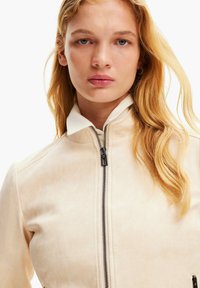 Giacca beige con una texture liscia, colletto alto e zip frontale. Presenta hardware della zip nera e colletto bianco a contrasto.