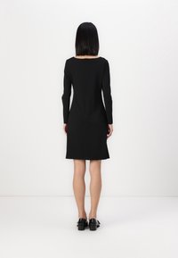 Černé šaty s dlouhými rukávy a rovnou siluetou, s hranatým výstřihem a zipem na zadní straně. Hladká látka, délka ke kolenům.
