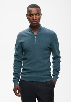 Homme portant un pull sarcelle à manches longues avec une fermeture éclair quart de longueur et un pantalon noir, debout avec une main dans la poche sur un fond blanc uni.