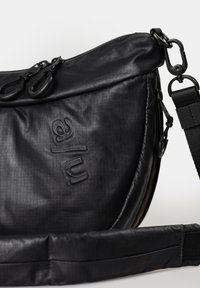 Black fabric bag with a curved shape, featuring a textured surface and embossed logo. Adjustable strap and zip closure with double pulls.