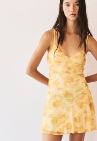 Gelbes Blumen-Minikleid mit tailliertem Oberteil, dünnen Trägern und einem fließenden Rock. Hergestellt aus leichtem, halbtransparentem Stoff mit einer weichen Textur.