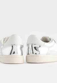Sneakers bianchi con una finitura lucida e riflettente. Presentano un tallone testurizzato e una suola in gomma color crema. Dettagli di cuciture a contrasto.