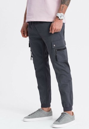 Mann trägt dunkelgraue Cargohose mit Klappen- und Reißverschlusstaschen, graue Sneakers, hellrosa Hemd und weiße Armbanduhr, steht auf weißem Boden.