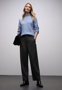 Pull côtelé bleu clair à col montant associé à un pantalon large gris foncé et des bottines noires, tenant une pochette noire.