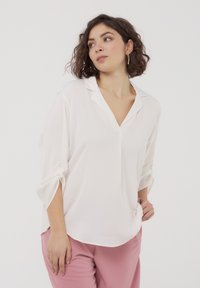 Blusa bianca con scollatura a V e maniche a tre quarti, con dettagli di bottoni sulle maniche, abbinata a pantaloni rosa. Texture liscia.