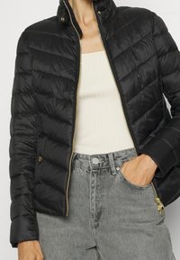 Černá péřová bunda s prošívaným vzorem, vysokým límcem a zlatými zipy. Kombinovaná s žebrovaným bílým topem a šedými džínami.