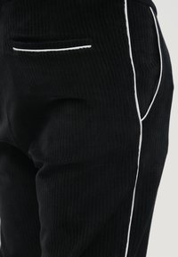 Pantalon en velours côtelé noir avec un liseré blanc le long du côté et une petite poche arrière à ouverture horizontale. Tissu texturé.