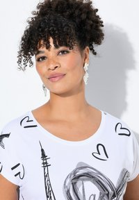 Camiseta blanca con diseños impresos en negro, incluyendo corazones y una Torre Eiffel. La modelo lleva pendientes en forma de lágrima brillantes y muestra su cabello rizado.
