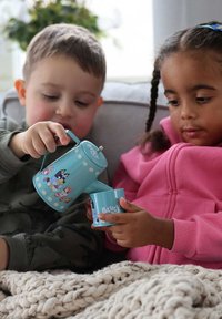 Lichtblauwe speelgoedtheepot met stippen en karakterontwerpen, gecombineerd met een bijpassend kopje. Kinderen zijn bezig met doe-ervaring op een bank.