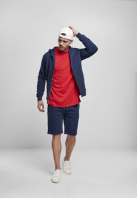 Marineblauwe hoodie, rode t-shirt, marineblauwe shorts en witte sneakers. De hoodie heeft een rits en een trekkoord, met een gladde textuur.