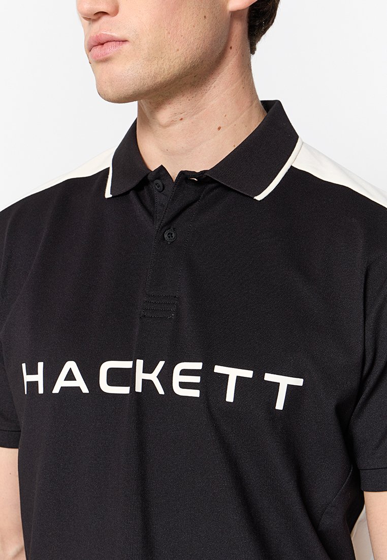 Schwarzes Poloshirt mit kontrastierendem weißen Kragen und Schulterakzenten. Auf der Vorderseite befindet sich ein auffälliges Textlogo "HACKETT". Strukturierter Stoff.