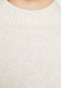 Pull en tricot beige clair avec un col montant côtelé, un tissu légèrement texturé et une apparence douce avec des variations subtiles de couleur.