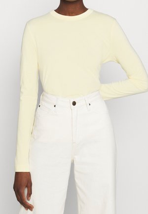 Personne portant une chemise à manches longues jaune pâle rentrée dans un jean blanc taille haute, un bras plié et la main sur la hanche.