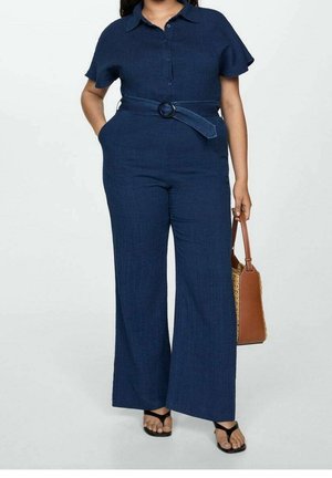Femme portant une combinaison bleu marine ceinturée à manches courtes, tenant un sac à main marron et portant des sandales noires.