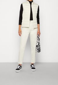 Gepolsterte elfenbeinfarbene Weste über einem schwarzen Langarmshirt, kombiniert mit hellen Hosen und schwarz-weißen Sneakern. Hält eine gemusterte Tasche.