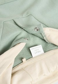 Hellgrünes Babybekleidungsstück mit weichem Material, Druckknopfverschlüssen und beiger Fütterung. Aufschrift mit "NEXT baby."