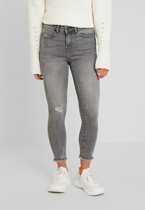 Femme portant un jean skinny gris clair usé avec des ourlets effilochés, un pull en maille blanc et des baskets blanches épaisses, debout sur un sol gris.