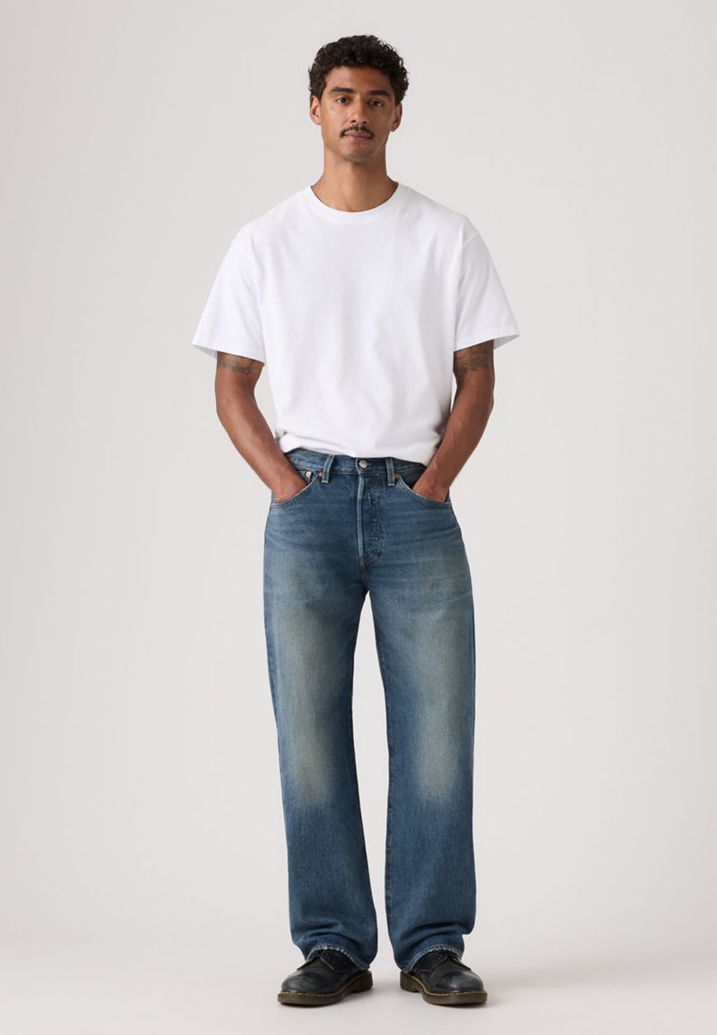 Camiseta blanca de manga corta combinada con vaqueros de mezclilla azul claro, de corte amplio, con lavado descolorido y diseño de pierna recta. Calzado negro completa el conjunto.