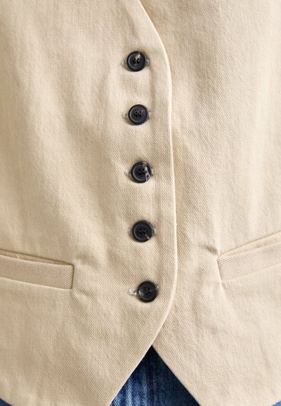 Beige vest van een textuurstof, met zes zwarte knopen aan de voorkant en twee kleine zijzakken. Het ontwerp is uitgevoerd met een spitse zoom.