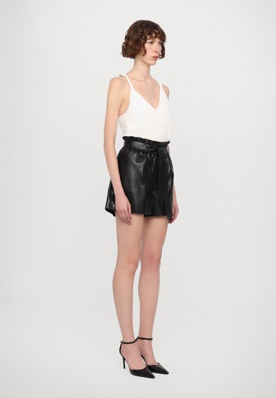 White sleeveless top with v-neck and strap details, paired with black leather high-waisted shorts and black strappy heels.