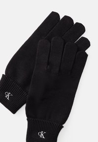 Luvas de malha pretas com cinco dedos, punhos canelados e um pequeno logo cK branco. Textura suave, design padrão de luva.