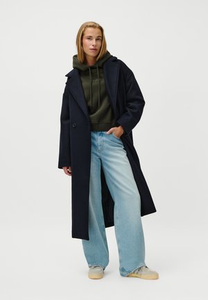 Navy long coat over a green hooded sweatshirt, paired with wide light blue jeans and gray sneakers, featuring subtle detailing and a relaxed fit.