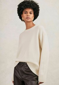 Tânără cu părul creț, purtând cercei de aur, un pulover oversized de culoare crem și pantaloni din piele maro închis, pe un fundal neutr.