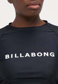 Čierny rash guard s krátkym rukávom so nápisom "BILLABONG" v bielej farbe cez hruď, vyrobený z hladkej a pružnej tkaniny.