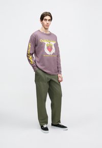 Purpurinė ilgomis rankovėmis marškinėliai su grafiniu piešiniu, vaizduojančiu rankas aplink širdį ir geltoną tekstą. Suteiktos su laisvomis alyvuogių žaliais kelnėmis ir juodais sportbačiais.